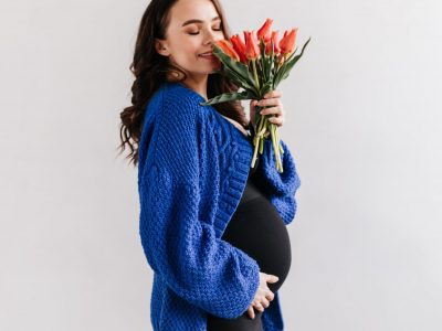 Happy brunette woman in blue cardigan and black dress smells tulips. Charming pregnant lady holds bouquet of flowers on isolated
